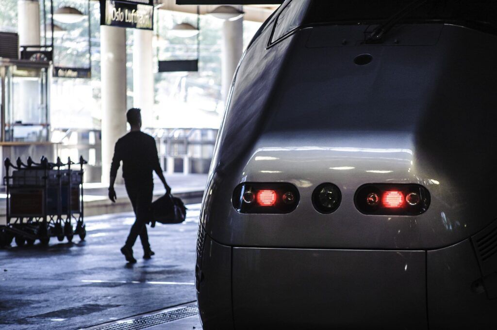 Et bilde av fronten på et tog ved en perrong. I bakgrunnen går en mann med en bag, og på et skilt i taket står "Oslo Lufthavn".