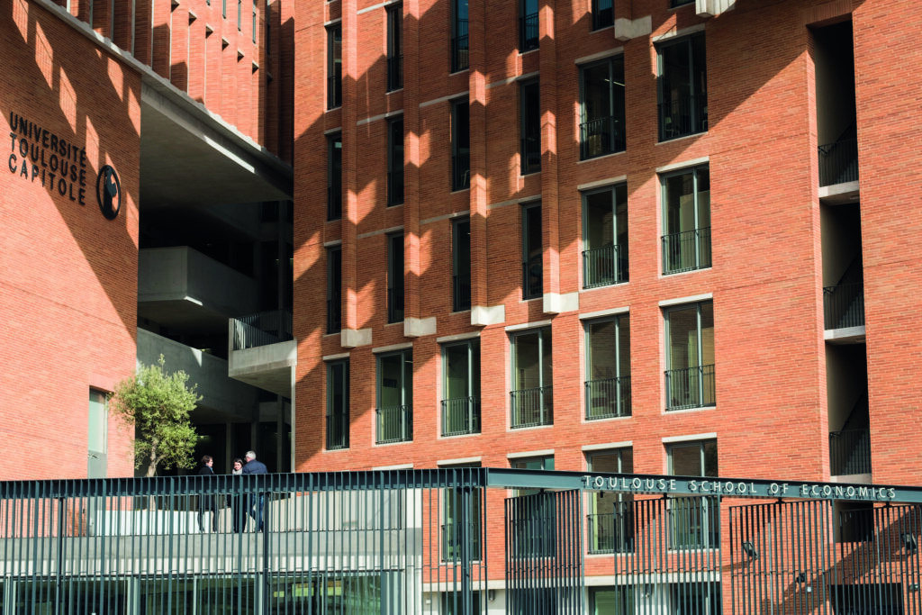 Moderne bygning i røde mursteiner tilhørende Université Toulouse Capitole, med skilt som viser "Toulouse School of Economics". Bildet viser store vinduer, skygger fra arkitekturen, og tre personer som står og snakker ved et rekkverk i forgrunnen.