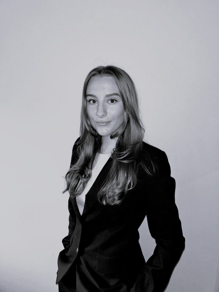 Portrettbilde av Jenny, som ser mot kamera, ikledd en sort dressjakke mot en hvit bakgrunn.