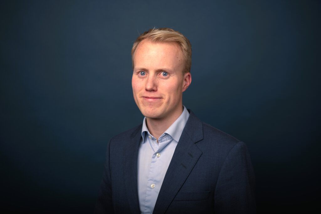 Portrettbilde av Johannes Hjartlie i lys blå skjorte og mørk blå dressjakke, foran en mørk blå bakgrunn.